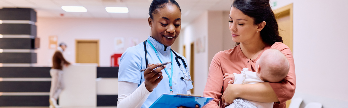 A medical professional looking at her notes with a woman holding a baby in a clinical setting.